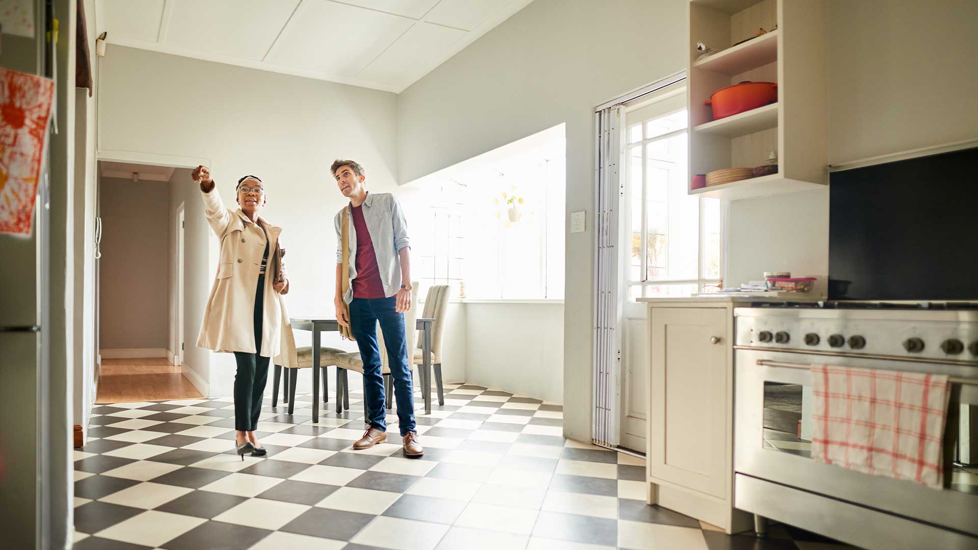 A home appraiser walks through a brightly-lit kitchen looking at fixtures with the home buyer