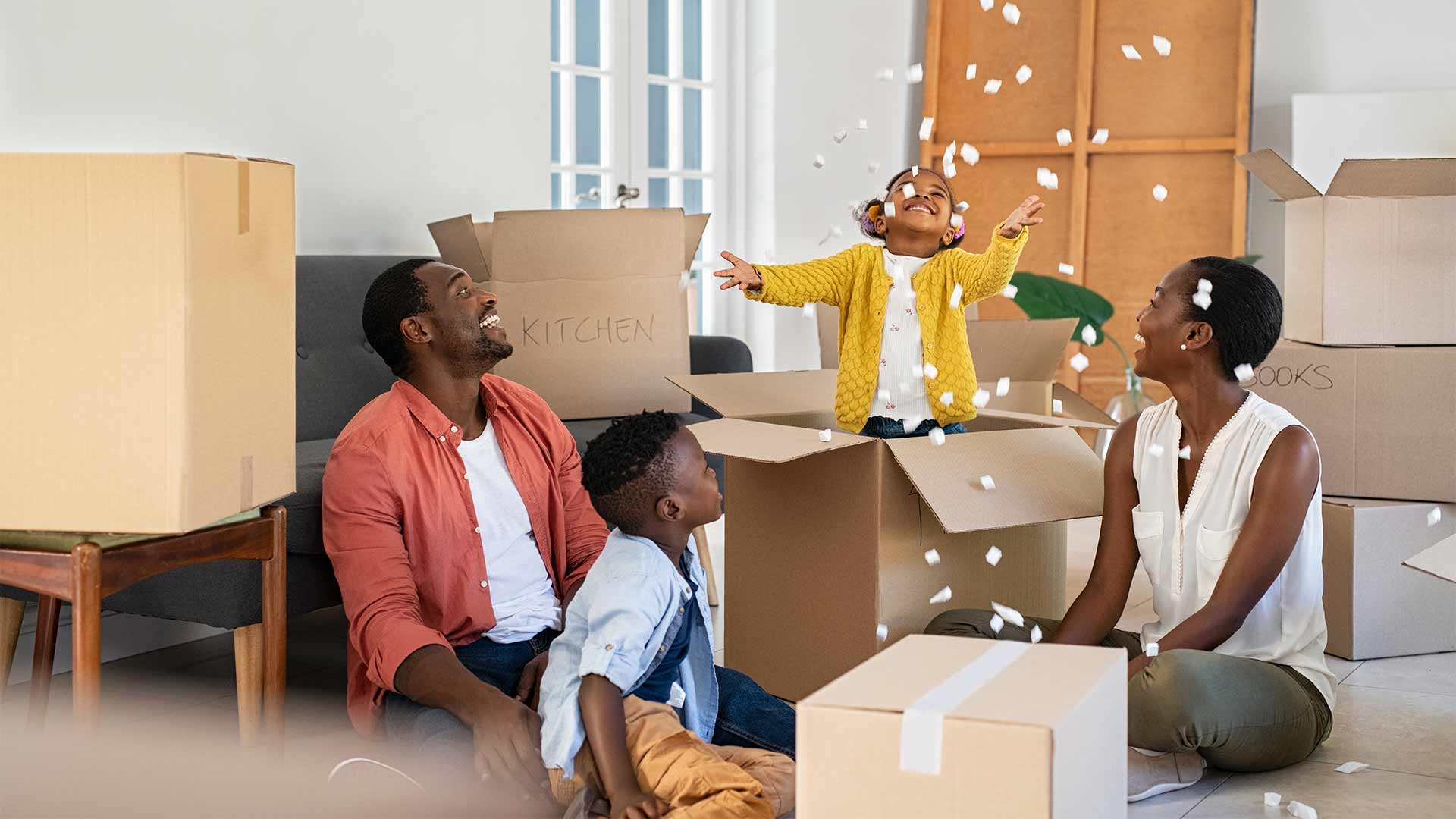A young family celebrates moving into their new home. Their daughter plays in a cardboard moving box in the living room