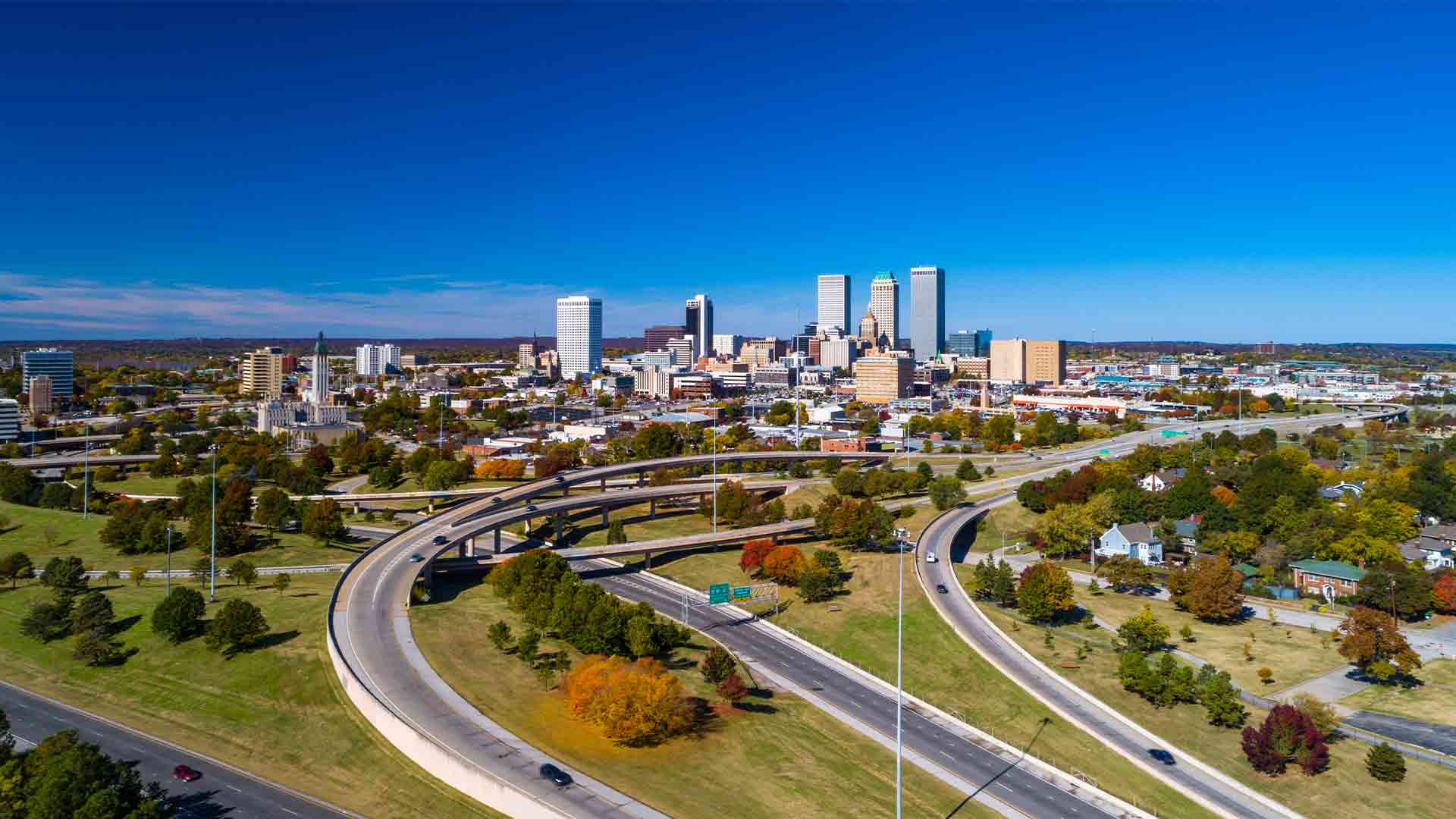 Aerial view of Downtown Tulsa skyline with grass, trees, and freeways in the foreground.